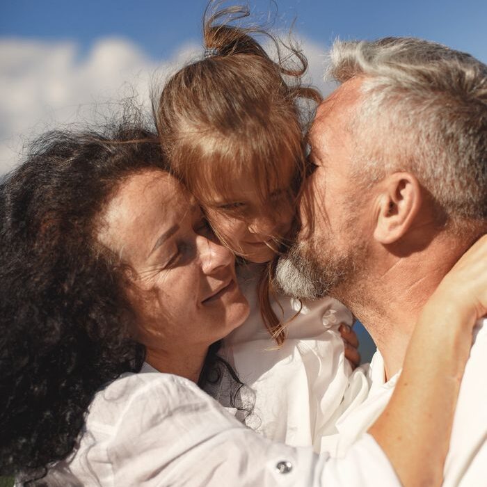 A heartfelt moment of grandparents hugging and kissing their granddaughter outdoors in bright daylight.
