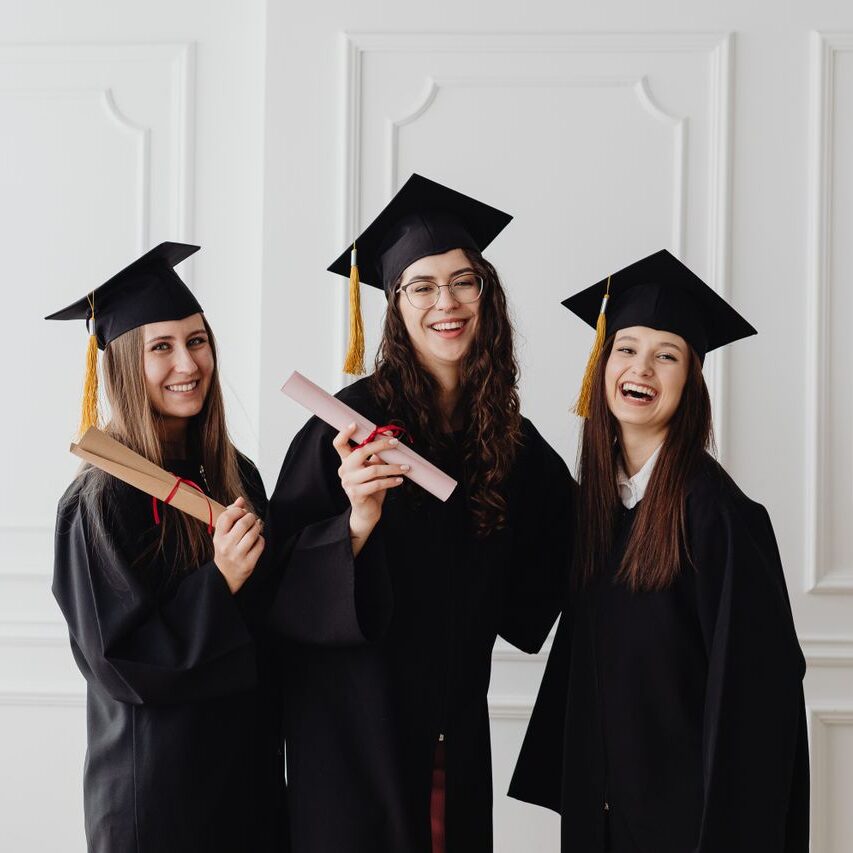 Three happy female graduates in caps and gowns holding diplomas and celebrating their achievement.
